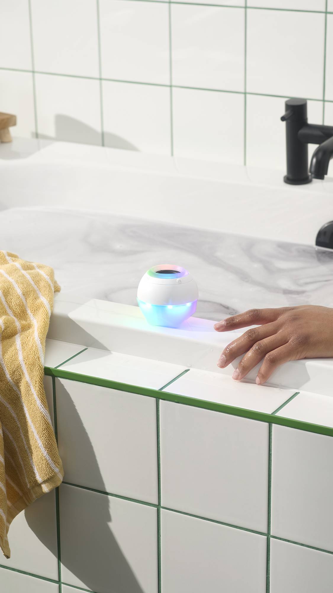 A round, white bath bomb-shaped waterproof speaker with glowing blue and rainbow lights sits on the edge of a white tiled bathtub filled with swirling bathwater. A hand rests on the tub nearby, next to a yellow striped towel.
