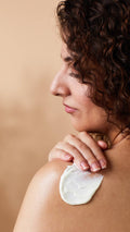 Close-up side profile of a person with dark curly hair smoothing a thick stroke of white cream onto their bare shoulder. Set against a warm, neutral beige background.
