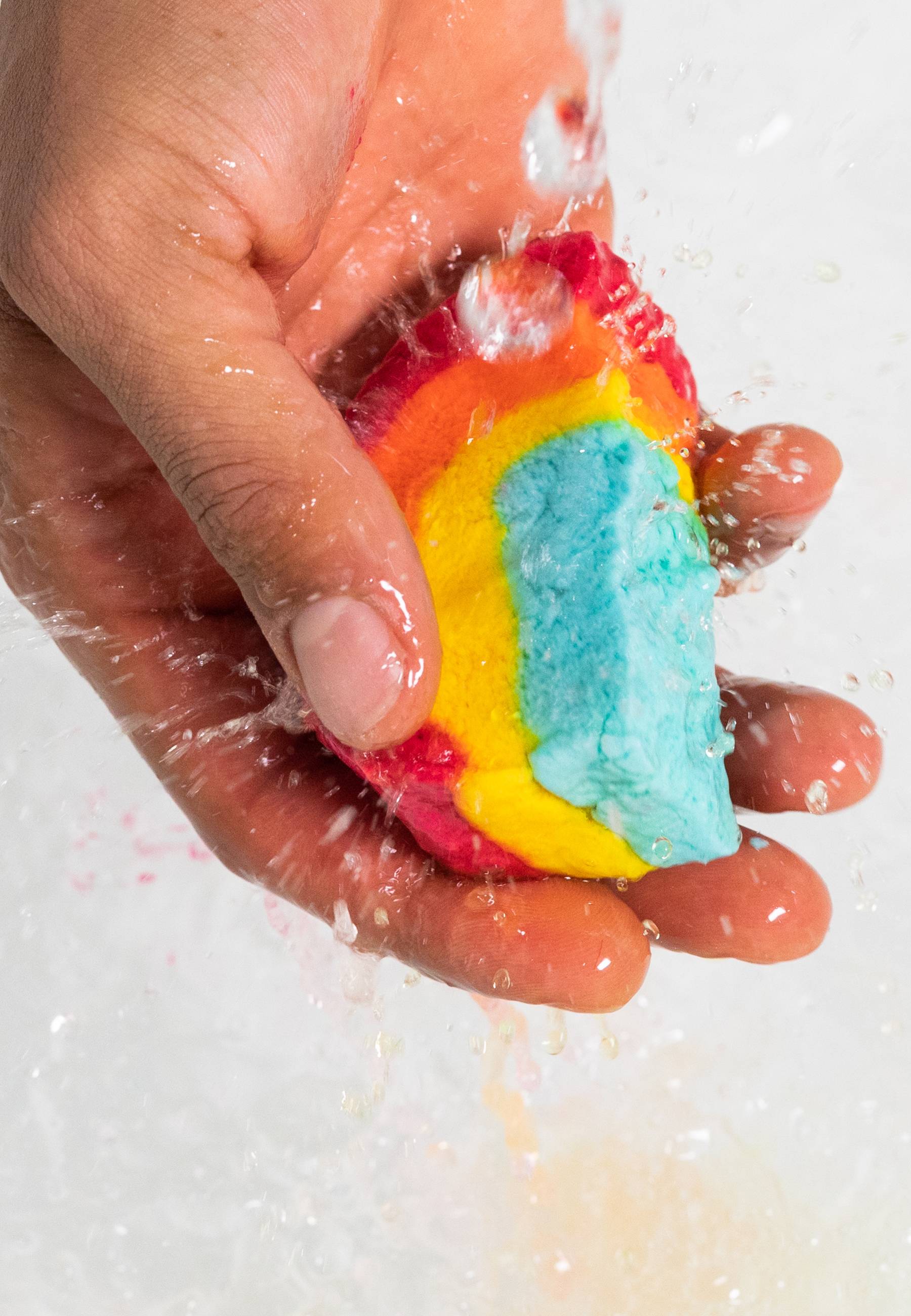 The image shows a light-toned hand vigorously squeezing and crumbling a 'Mini Rainbow' bubble bar. A strong stream of water splashes onto the bar, causing its colourful layers to dissolve into thick, bubbly foam in the bathwater below.