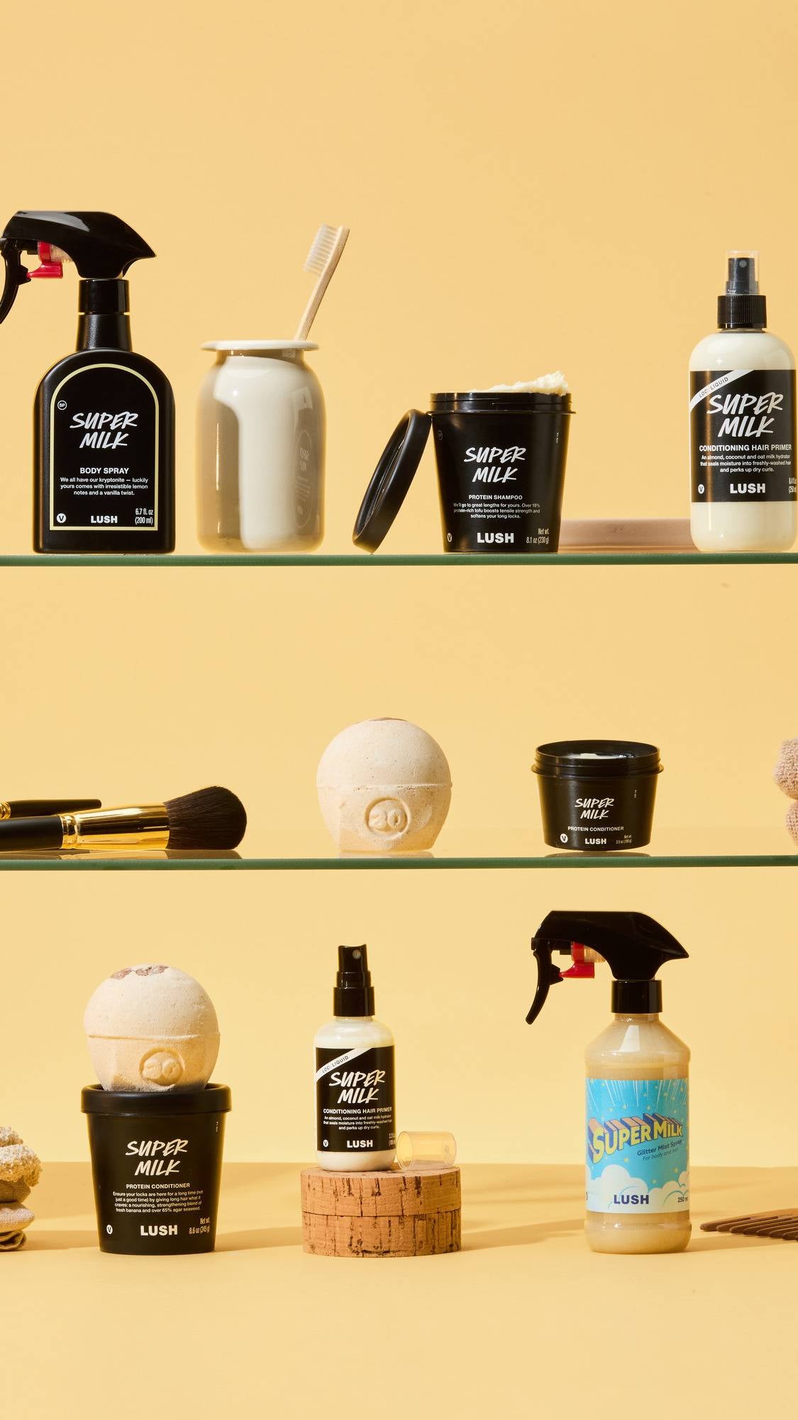 The Super Milk collection is arranged on glass shelves against a yellow background. It features the Body Spray, Hair Primer, Protein Shampoo, Conditioner, and Glitter Mist Spray in black and white packaging.