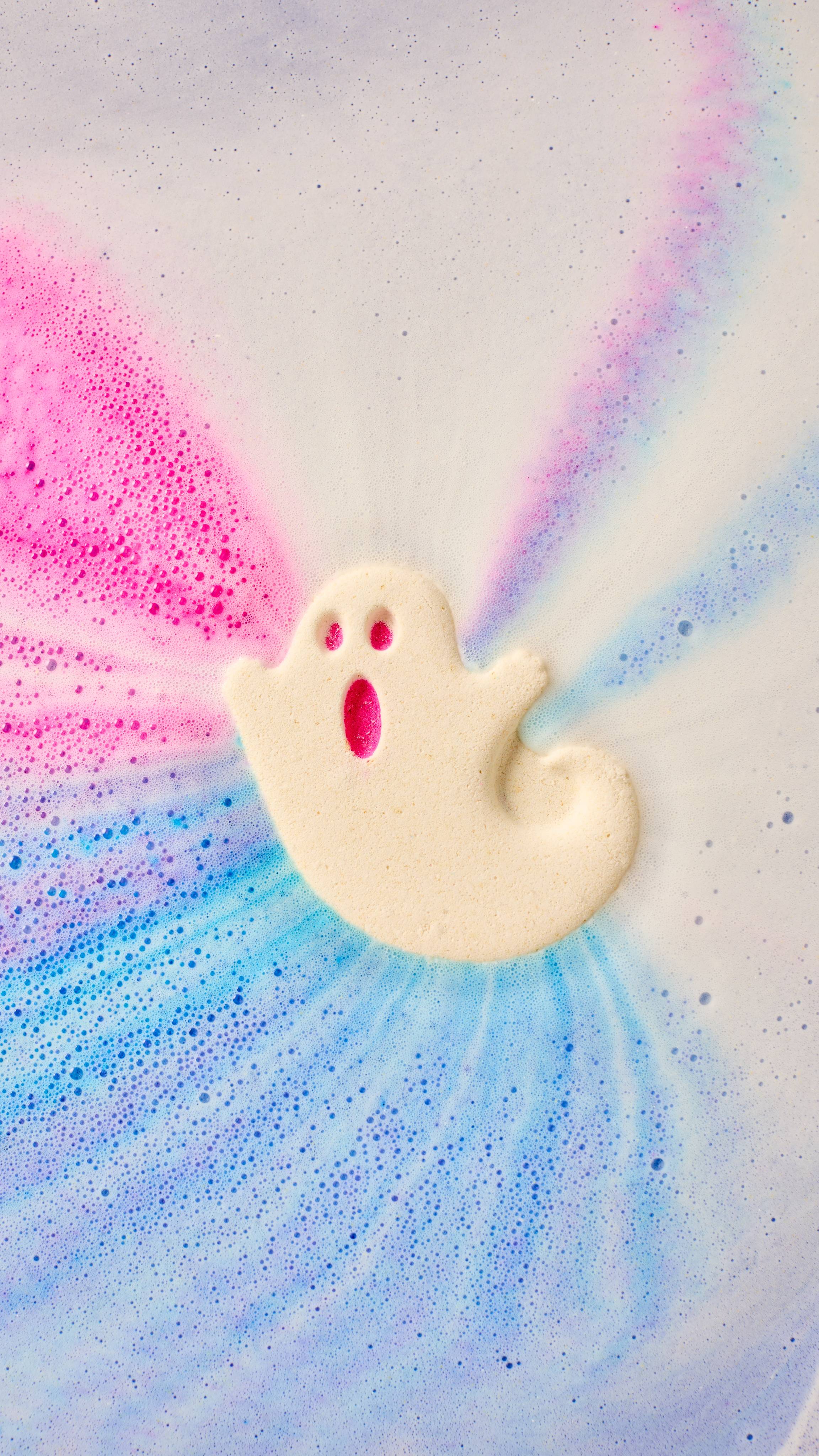 A cream-coloured ghost-shaped bath bomb dissolves in water, releasing vibrant trails of pink, purple, and blue foam. The ghost has a surprised expression with hot pink hollows for its eyes and mouth.