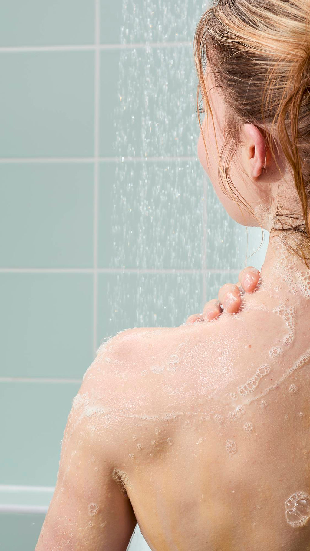 A woman seen from behind in the shower, applying a light, bubbly lather to her shoulder. The background features light mint green wall tiles and the soft, vertical spray of falling water.