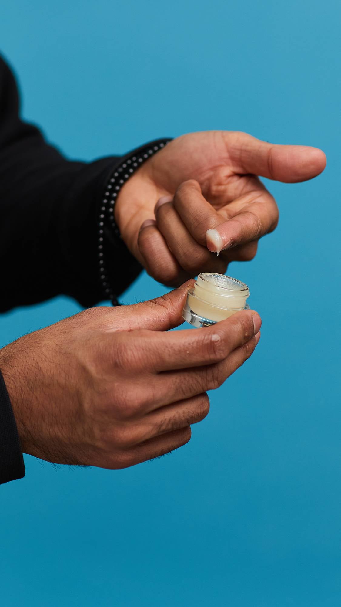 A close-up shot of the model's hands on a sky-blue background. They are holding a small, glass pot as they swipe the Shimmy Shimmy lip gloss onto the tip of a finger.